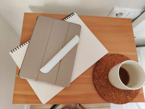 Closed tablet with stylus on notebook and cup of coffee on wooden bedside table. Minimalist home office workspace. Interior design concept for remote student or freelancer lifestyle.