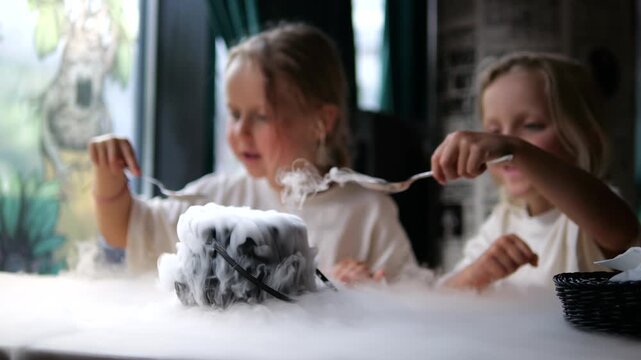 Two excited little girls sitting at a restaurant table are mesmerized by a dessert served in a cauldron, playfully touching the thick white smoke created by the sublimation of dry ice