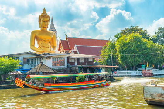 Traditional Thai long tail boat moving along canal in Bangkok with giant golden Buddha statue at Wat Paknam Phasi Charoen, Thailand. Urban waterway scene combining transport and religious architecture
