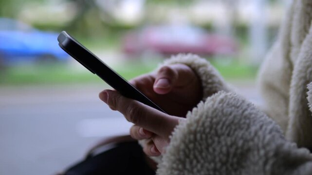 Close up view of a young woman's hands in a cozy fleece jacket typing and scrolling on a modern smartphone, with a softly blurred city street providing the background for this communication concept