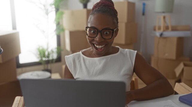 Young woman pumps fists and smiles widely at laptop surrounded by stacked moving boxes in a building; joy success.
