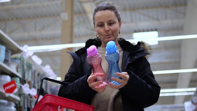 Thoughtful female customer comparing two colorful reusable water bottles, a pink and a blue one, while shopping for eco friendly products in a large department store aisle