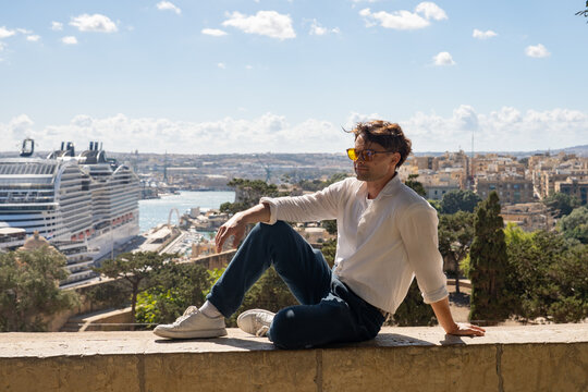 Young man in sunglasses sitting on a historic wall with a giant cruise ship in the background. Mediterranean travel concept in Valletta, Malta.