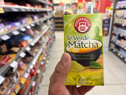 Alicante, Spain - April 25, 2026: Hand holding Pompadour green tea with matcha box in a grocery store aisle filled with various packaged products and snacks
