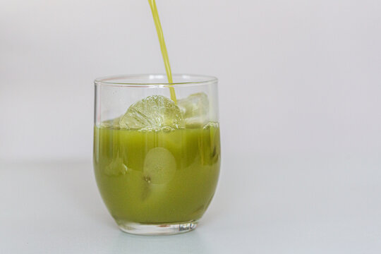 Green matcha beverage being poured into a clear glass with ice cubes and a slice of lime, set against a light background for a refreshing drink presentation