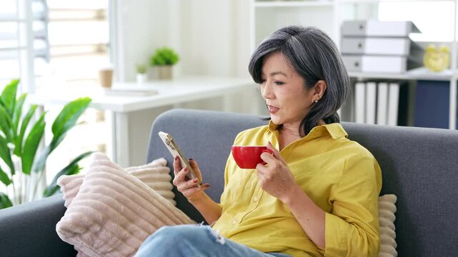 Happy middle aged asian woman using smartphone sitting on couch at home. holding cellphone browsing internet, texting messages on mobile cell phone technology.