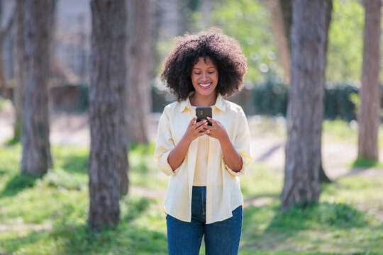Young African woman uses smartphone for texting while standing among trees in a park on a sunny day