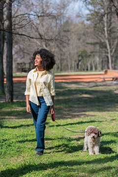 African woman walks dog in the park on a sunny day surrounded by trees and grass