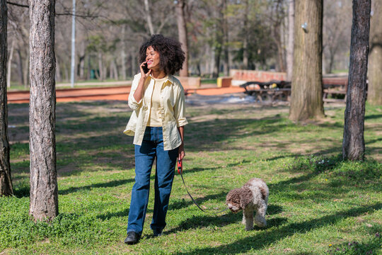 Young African woman walks dog in the park on a sunny day surrounded by trees and grass