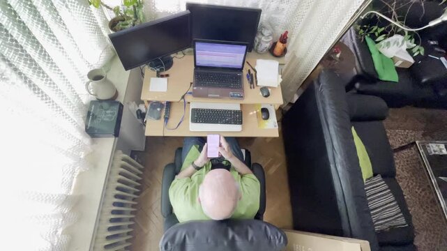 Viejo pensionista activo con m&oacute;vil, ordenador port&aacute;til y pantallas sentado en silla de despacho oficina de casa visto desde arriba