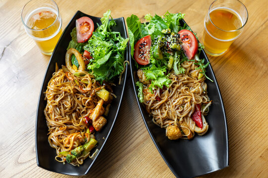 Asian noodle dishes with salad and beer. Top view of two Asian noodle plates with fresh salad, tomatoes, vegetables, seafood, and beer on a wooden table.