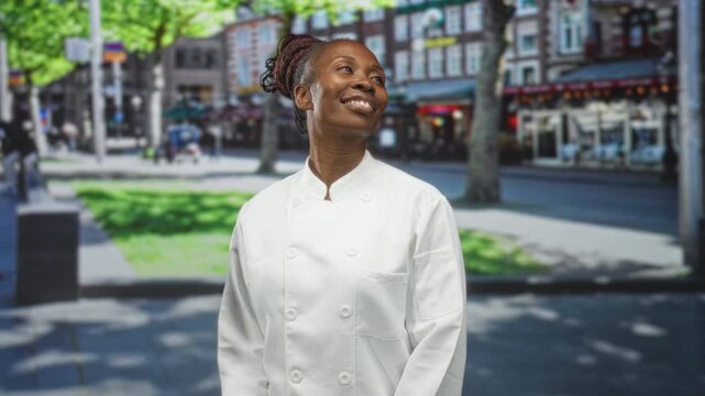 Young african american cook in white chef coat looks up smiling, face turned skyward on a street; pride aspiration.