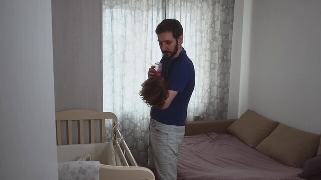 Stay-at-home dad bottle feeding toddler in nursery. Single father doing daily childcare routine, standing and holding his little girl while she drinks formula from bottle