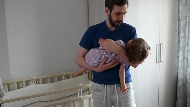 Sleepy toddler girl being rocked by caring father near wooden crib. Child comfort and security moment showing little girl drifting to sleep in dad's arms in cozy nursery