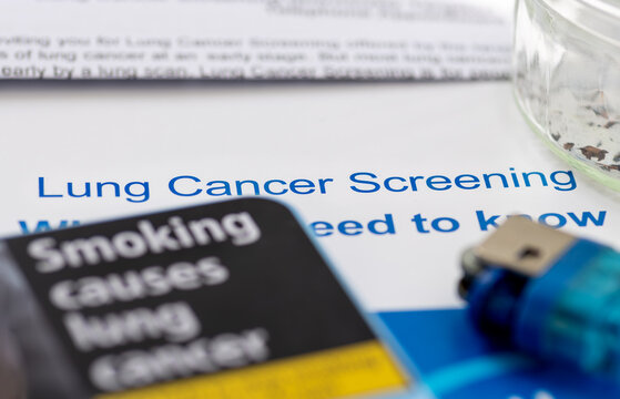A macro close up of a cigarette case with a health risk warning, a lighter and ashtray on a letter and information leaflet on lung cancer screening.