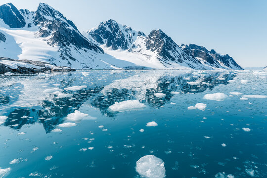 Svalbard, Svalbard and Jan Mayen - 24 April 2026: View of snow-capped mountains reflecting in the calm blue waters of a fjord filled with floating ice chunks under a clear sky.