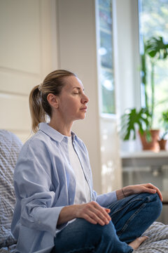 Serene mature woman practicing deep breathing and mindfulness exercises, relaxed female comfortably sitting on sofa cross-legged concentrating on breath, physical health and mental clarity rituals.
