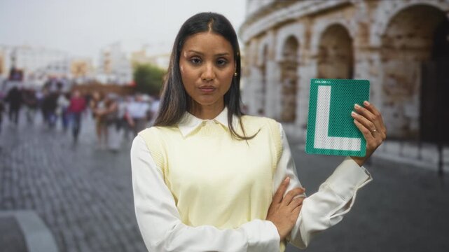 Young brunette woman holds green learner l sign with right hand and crosses left arm on cobblestone street by ancient building; confidence learning.