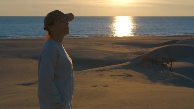 Silhouette of a woman watching the sunset over the sea. Side view of a thoughtful woman in a cap standing on sandy dunes, peacefully contemplating the beautiful golden sunset reflecting on the ocean