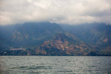 Misty mountains loom over calm lake waters. Green slopes rise from the shoreline's edge. Clouds drift over rugged, rocky peaks. Faint settlement lines the distant shore © The Adaptive