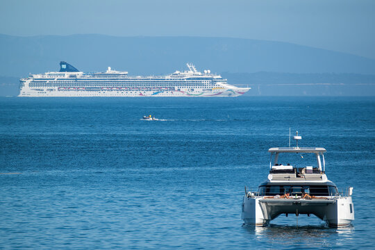 Cascais, Portugal - 18 April 2026: View of a large cruise ship sailing in the distance and a modern catamaran with people on deck in the foreground on the calm blue sea.