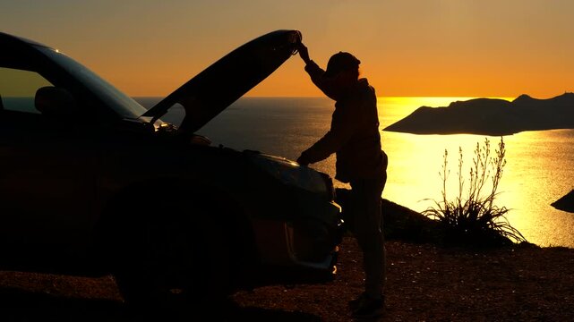 Silhouette of a man closing the car hood at sunset. Silhouetted man closing the hood of his car after an inspection, standing on a cliff overlooking the sea during a beautiful golden sunset