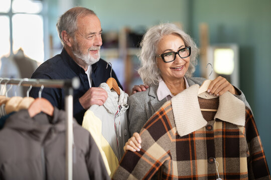 Senior wife with husband client pre-examine fabric and style of product, is busy choosing down jacket or drap coat. Client enjoys shopping, clothing and accessories for whole family, seasonal sale