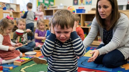Naklejka premium Upset little boy covering his ears and crying in a colorful preschool classroom while a caring teacher tries to comfort him