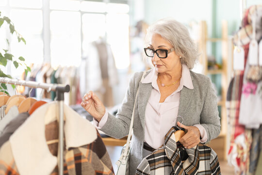 Senior woman client pre-examine fabric and style of product, is busy choosing down jacket or drap coat. Client enjoys shopping, clothing and accessories for whole family, seasonal sale