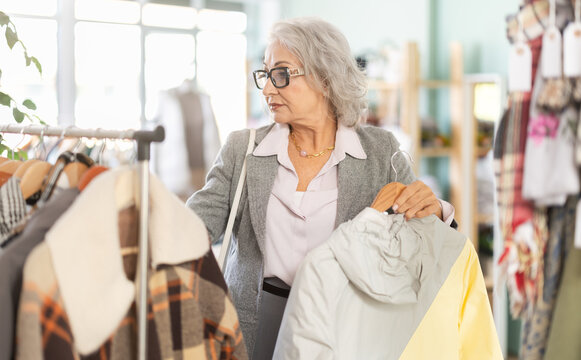 Senior woman client pre-examine fabric and style of product, is busy choosing down jacket or drap coat. Client enjoys shopping, clothing and accessories for whole family, seasonal sale