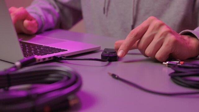 Preparing audio setup with focus on connection and ambient lighting. Attaching audio cable to laptop emphasizing precise finger placement and purple studio illumination
