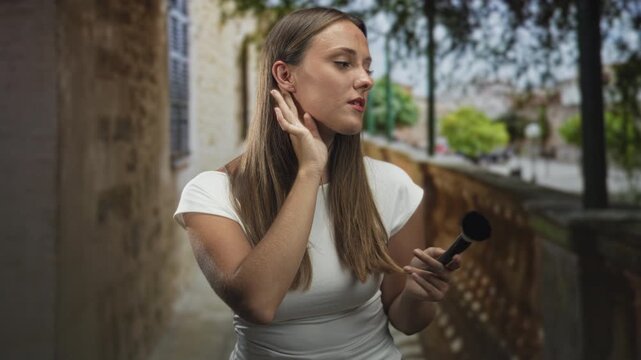 Young woman holds a makeup brush and touches her ear while checking her face beside a stone balustrade on a tree lined street; confidence beauty routine.