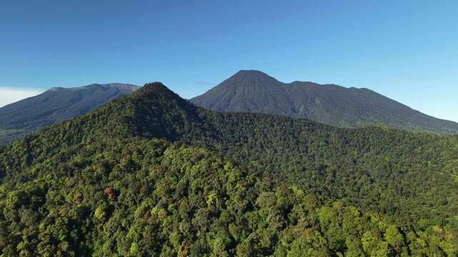 A drone flies over green forest hills with towering mountains in the background under a clear blue sky. Mount Gede Pangrango National Park, Bogor.