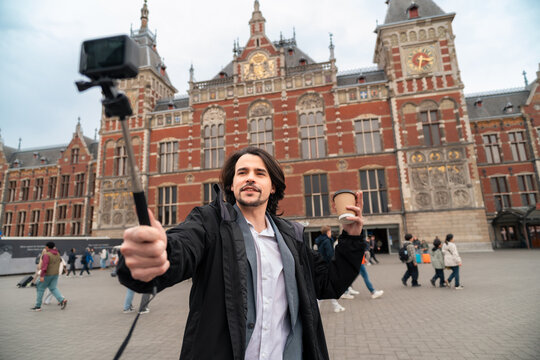 Vlogger recording travel content in amsterdam, holding coffee and selfie stick in front of central station