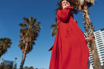 Obraz premium Young woman in a red coat smiles joyfully while walking outdoors on a sunny day with palm trees and modern buildings in the background.