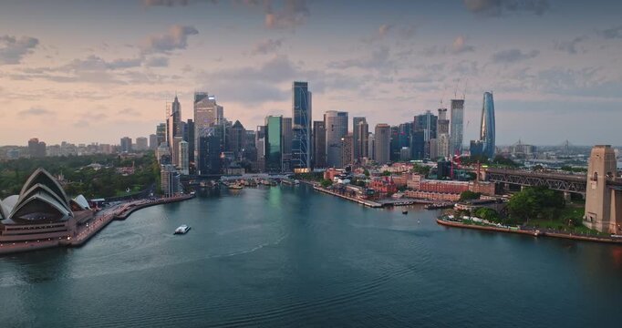 Panoramic view of Sydney sunset cityscape with iconic landmarks - the Opera House and Harbour Bridge, bathed in the soft light of dusk with city skyscrapers in the background. Aerial view drone flight