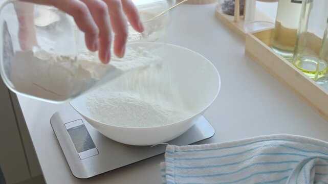 Hand pours flour into bowl on kitchen scale during baking preparation. Concept of dough making, ingredient measuring and homemade cooking