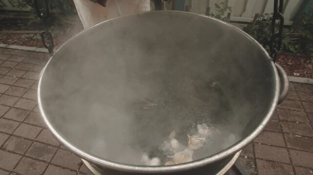 Steaming pot with hands adding, dried herb bundle lowered into boiling water, smoky vapor rising, outdoor stove on patio, simmering infusion, closeup on palms and rustic cookware
