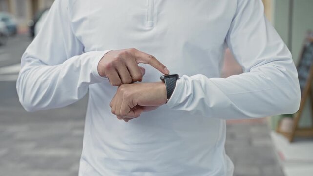 Young hispanic man in white long sleeve points finger to smartwatch on wrist while standing on a city street sidewalk near storefront; time pressure urgency.