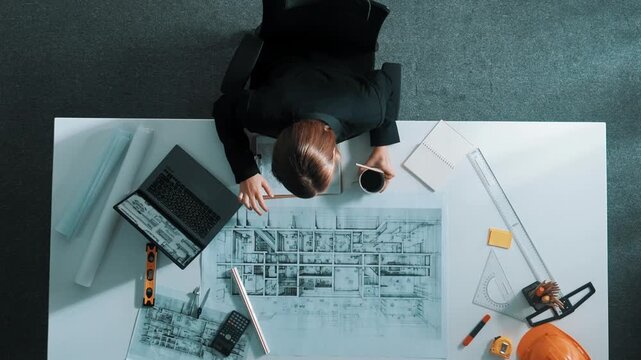 Aerial view of architect drawing while working at laptop. Top down view of caucasian engineer designing blueprint and drafting house model at table with equipment and safety helmet. Alimentation.