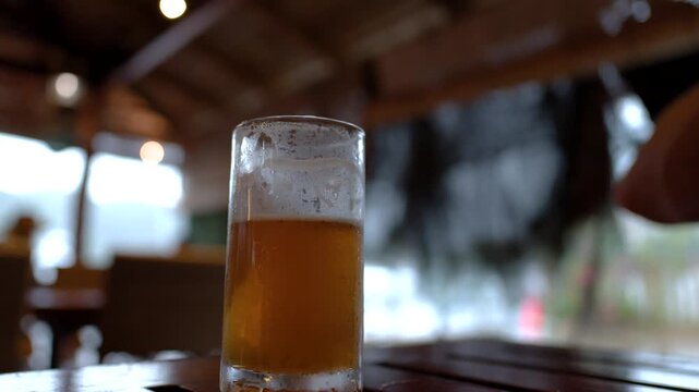 Two glasses of golden beer on a rustic wooden table in a well-lit indoor setting, one glass is picked up and placed down.