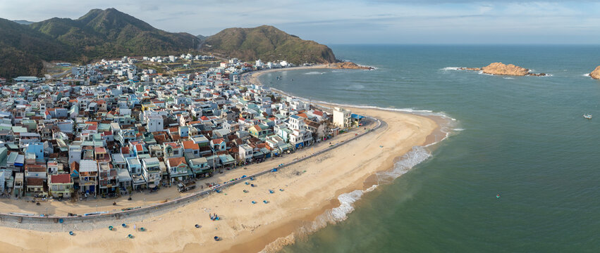 A panoramic view of a coastal town of Nhon Hai, Quy Nhon, Binh Dinh, Vietnam, with fishing boats on the water and mountains in the background.