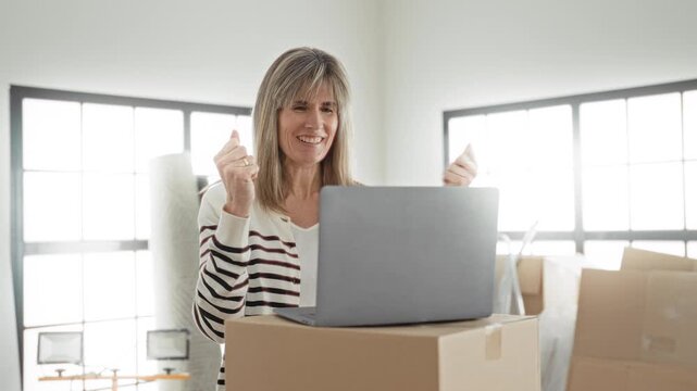 Woman smiling with laptop and moving boxes raises fists in celebration in a bright building; small business joy.