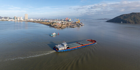 Fototapeta premium A cargo barge navigates the harbour in Quy Nhon, Binh Dinh, Vietnam. The busy port with cranes and ships is visible in the background.