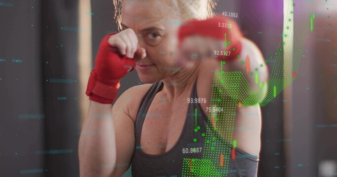 Punching female boxer practicing punch in studio, wearing dark tank, red wraps, candlestick chart