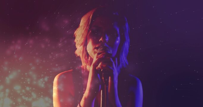 Singing singer wearing sleeveless top, gripping mic and stand on stage with lights, haze particles