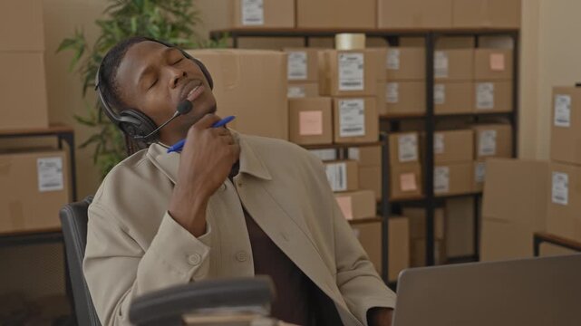 Man wearing headset holds pen to chin and leans back by laptop and stacked parcels on metal shelving in a warehouse shipping office, surrounded by boxes; calm focus.
