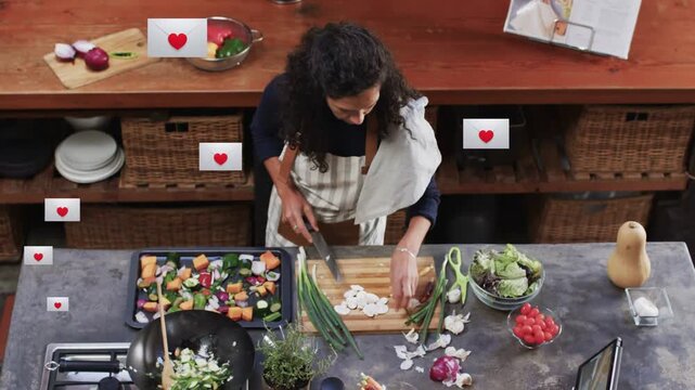 Woman chopping, stirring at prep island, tapping tablet, causing hearts to follow recipe cues