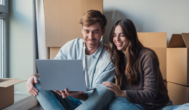 Smiling couple sits on the floor among cardboard boxes, sharing a laptop as they unpack and plan their new home, enjoying a relaxed moment of togetherness and excitement about moving in.