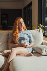 Obraz premium Relaxed redhead woman sits on a light sofa in a bright living room, holding a mug of coffee and enjoying warm morning sunlight in casual loungewear for a quiet, cozy moment.